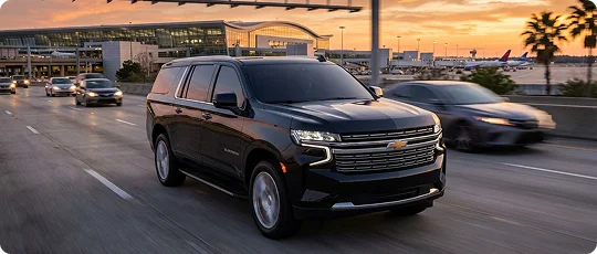 A black Cadillac Escalade drives along an airport access road with terminals and palm trees in the background at sunset.