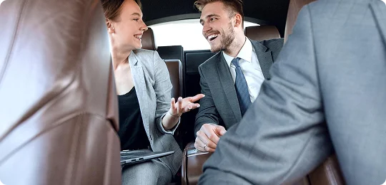 Businesswoman and businessman in gray suits having a cheerful discussion inside a luxury car’s brown leather backseat during a professional ride.