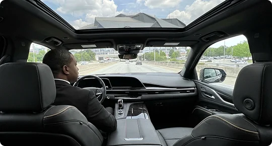An interior view from the backseat of a luxury SUV, showing a chauffeur in a black suit driving along a highway. The vehicle features a large panoramic sunroof revealing a cloudy sky and the Mercedes-Benz Stadium in the distance. The cabin is finished with premium black leather and wood trim.