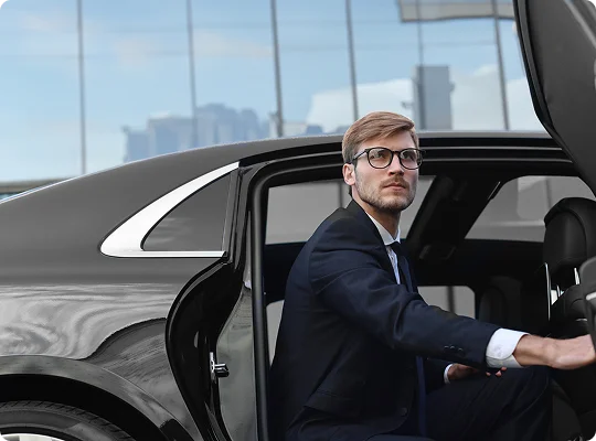 Blonde businessman in glasses stepping into a sleek black car with a chrome-trimmed door, seen from a close-up side angle.