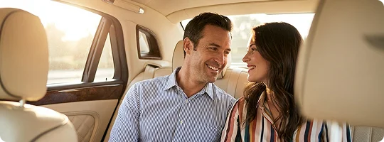 A decent couple smiles at each other while comfortably seated in the back of a luxury car with plush white leather interior.
