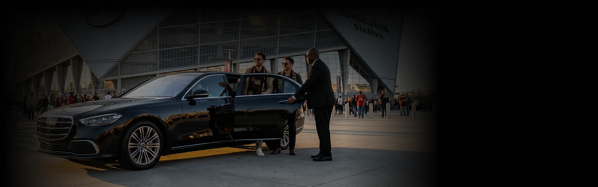 two young football fans stepping out of a black business sedan (Mercedes-Benz S-Class) parked outside Mercedes-Benz Stadium, Atlanta Georgia. Chauffeur assists them by holding the rear door open and the background scene shows, stadium building, fans crowd and sunlight.