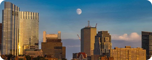 A panoramic skyline view of Downtown Atlanta city buildings under a daytime sky, with the moon faintly visible above.